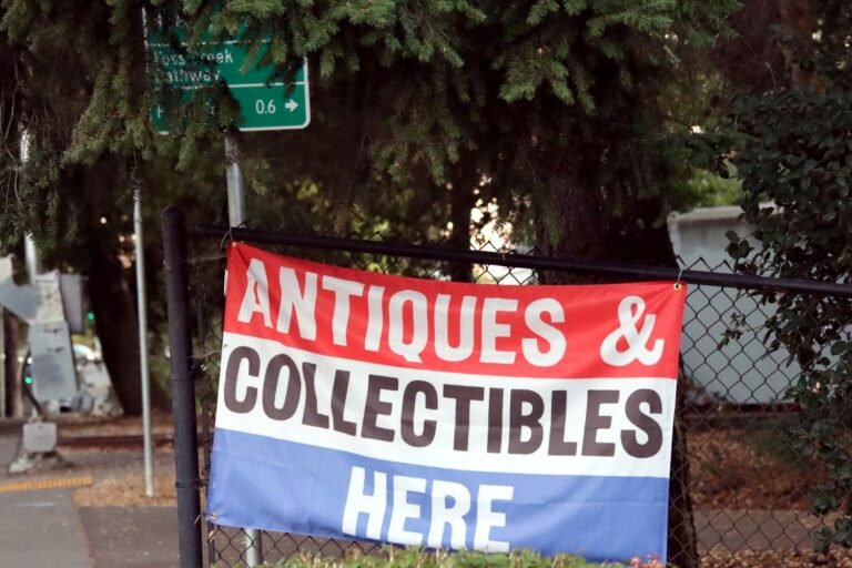 An "Antiques & Collectibles Here" banner on a fence beside a Foss Creek Pathway sign in Healdsburg